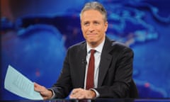 Middle-aged white man with white-gray hair, black suit, white shirt, red tie, sitting at TV desk in front of blue background holding powder blue papers.