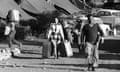 A woman in a dress carrying two suitcases walks on a dirt road past tents, cars and a man with a box on his shoulders in the sun in Cyprus in 1974