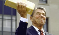 a man in a suit and tie holds up a large block of cheese