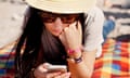 young woman text messaging on mobile phone while lying on a colourful checked red, orange, yellow and blue picnic blanket on a beach: she has long dark hair and wears large sunglasses, jeans and a pale grey T-shirt