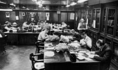 Two large tables filled with people on both sides in the corner of a room with panelling and built-in bookcases, with huge bundles of paper stuck on spikes in between every occupant