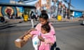 A young Latina woman with black hair pulled into a bun on top of her head, smiling and wearing a pink sweatshirt, holds an infant girl, also in pink, and a long paper box covered in cellophane filled with glazed puffy doughnuts. Behind them is a one-story building with a peaked roof, covered in a colorful mural with religious themes.