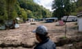 A white man is seen from the back, blurry in the foreground, wearing a black jacket, baseball hat and glasses, as he surveys a wash between two stands of twos, with damaged homes, vehicles and land caked in mud.