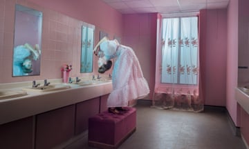 A sterile pink-tiled bathroom, with a girl bent right over backwards to see herself in the mirror (her feet face away from the mirror)