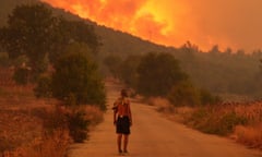 A man is seen from behind as he stands on a road looking at a wildfire. The air is smoky and the sky is orange