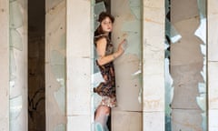 A barefoot young woman stands amid broken glass in a window frame