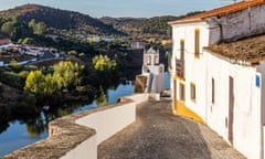 Buildings on a walled, cobbled street in Mertola, with a river on the left and hills behind it