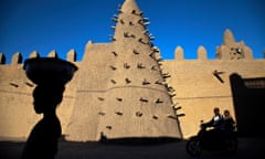 A woman in silhouette passes the minaret of a mosque made out of mud reinforced with sticks