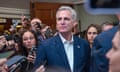 A middle-aged white man with white hair, wearing a blue suit and no tie, stands tall amid a scrum of reporters, looking calmly over their heads.