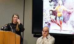 A woman stands at a podium wiping a tear from her cheek as a man sits next to her, his hand over his mouth.