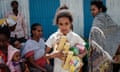 A girl holds food items and looks at the camera as women look on behind her.