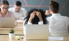 Frustrated woman feeling despair in a shared office, holding her head in her hands over her laptop.