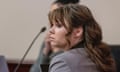 A young, white-presenting woman with long brown hair and bangs, wearing a gray blazer, is seen from the back sitting at a courtroom table and looking to her left as if listening to someone speaking.