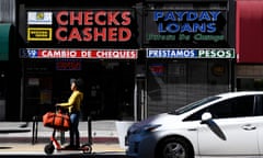 On a sunny urban street, a person rides a scooter past two storefronts, one with a red sign that says 'Checks Cashed' and the other with a blue sign that says 'Payday Loans'.