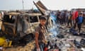 A boy with cuts on his face and a distressed look on his face stands in front of a burned car and debris from the explosion at the camp