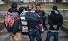 FLINT IS A PLACE<br>An officer holds a small child while her father is being searched and questioned.