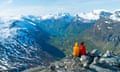 Two people sit on a rocky outcrop overlooking a range of snow-capped mountains