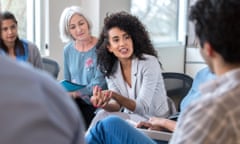 A cheerful young businesswoman sits in a circle during a staff meeting. She puts her hands together as she speaks to an unrecognizable coworker.