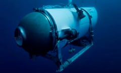 An underwater picture of different shades of dark and light blue, with light from above shining on a metal capsule shaped like a pill with a porthole on the end facing toward the camera.