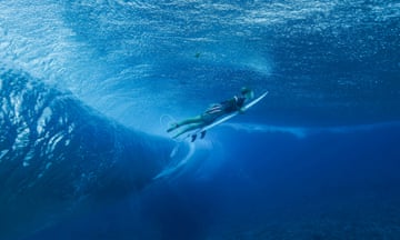 A surfer underwater holding his surfboard, heading upwards to break the surface of the water