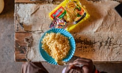 A hand holding a spoon above a bowl of instant noddles next to its colourful packet on a wooden table