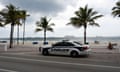 Police car alongside flat, narrow stretch of beach on the other side of a flat, brick walkway, with palm trees.