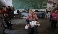 A Palestinian woman in a salmon pink headscarf sits in a classroom with a swaddled  baby on her lap. Around her are older children. Behind her the blackboard has chalk writing on it.