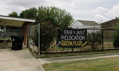 A hand-painted sign hangs from a black iron fence around a white home that reads 'Fair and just relocation for Gordon Plaza residents'. A dog runs across the carport to the left.