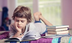 little boy lying on bed reading next to stack of books