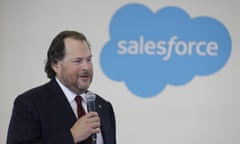 Larger white man with long brown hair, light beard, and suit hold microphone in front of blue, cloud Salesforce logo on white wall behind him.