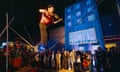 A performer plays the violin on a tightrope, watched by a crowd in front of a looming building labelled ‘eviction’