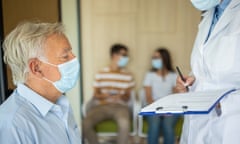 A doctor is assisting a senior man to fill medical consent form while he's in a waiting room in a medical clinic, they are wearing face masks
