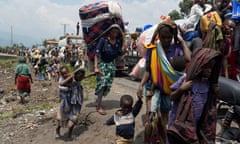 People carrying belongings and children walk along a road beside trucks