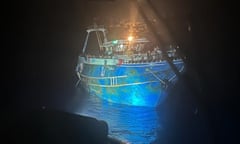 The Adriana, an ageing fishing trawler, is seen illuminated in the dark at a port with people crammed onto its decks and even onto the roof of its cabin. It is painted blue with large patches of rust on its hull.