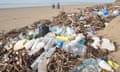 driftwood, lots of plastic bottles and other pollution on beach, with two figures on bikes in background