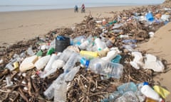 driftwood, lots of plastic bottles and other pollution on beach, with two figures on bikes in background