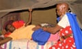 An elderly woman with shaved head and beaded earrings feels the bump of a pregnant woman lying on a bed