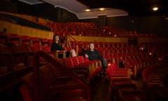 Woman and man sit on bar stools in front of chandeliers and red curtains