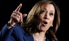 Black and South Asian woman with shoulder-length brown hair and blue pantsuit and pearls speaks and gestures with one finger on a stage.