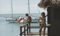 A couple on a porch in the Bahamas in 1967, she posing in a bikini, he sitting on the edge fishing