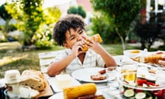 Young Boy Eating Barbecued Corn On The Cob<br>Posed by model A young boy with curly hair sitting at a table in his back garden and eating a barbecued corn on the cob.