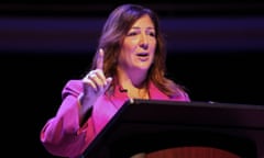 A woman dressed in pink at a lectern raises one finger in the air.