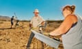 Three white people under a blue, cloudless sky stand on a sandy, barren landscape working with shovels and metal tools.