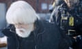 An elderly man with white hair and a beard being led in handcuffs by an official