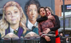 Two women, one tied with rope, embracing in front of a mural of the Derry Girls