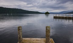 A view across lake Windermere on a dark, cloudy day, showing two wooden ramps , with mountains and sailing boats in the background.