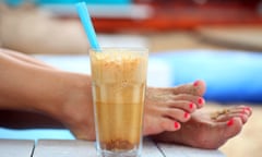 A glass of iced coffee with a straw placed on wooden planks. Women's sandy feet in the background