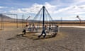 A wide shot of an old-fashioned merry-go-round, with cords attached from a center pole to the round edge, and five or six young people pushing it quickly, on a rocky flat yard with swings and a basketball hoop, under a huge blue sky, with low mountains beyond.