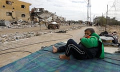 A young boy lies on a blanket with a donkey and cart in the background in front of a bombed building