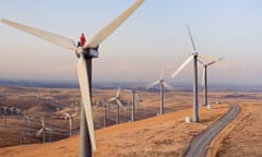 Worker standing on wind turbine at wind farm<br>High angle view of a male worker standing atop a wind turbine at dusk wearing protective work clothes and safety hardhat inspecting machinery